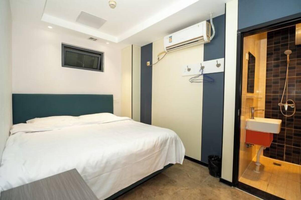 Habitación de hotel con cama, equipo de aire acondicionado, ventana y baño con ducha y lavamanos.