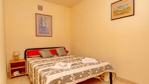 Een net opgemaakt bed met een beddegoed met een patroon, een nachtkastje met een lamp en twee schilderijen aan de muur.