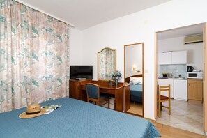 Habitación de hotel con cama, escritorio con televisor, espejo, silla y área de cocina con microondas, además de una mesa de comedor.
