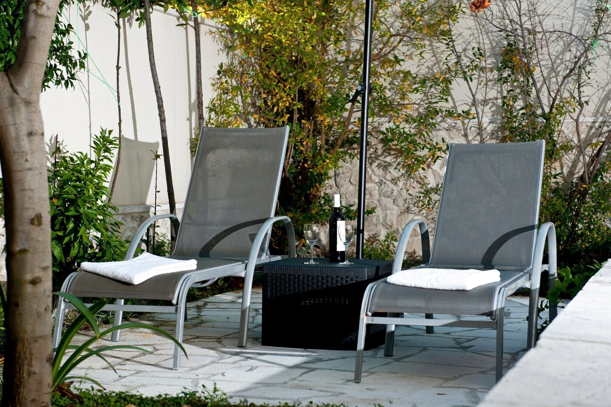 Two metal chairs with cushions, a black table, and a bottle of wine on a patio with stone flooring and greenery.