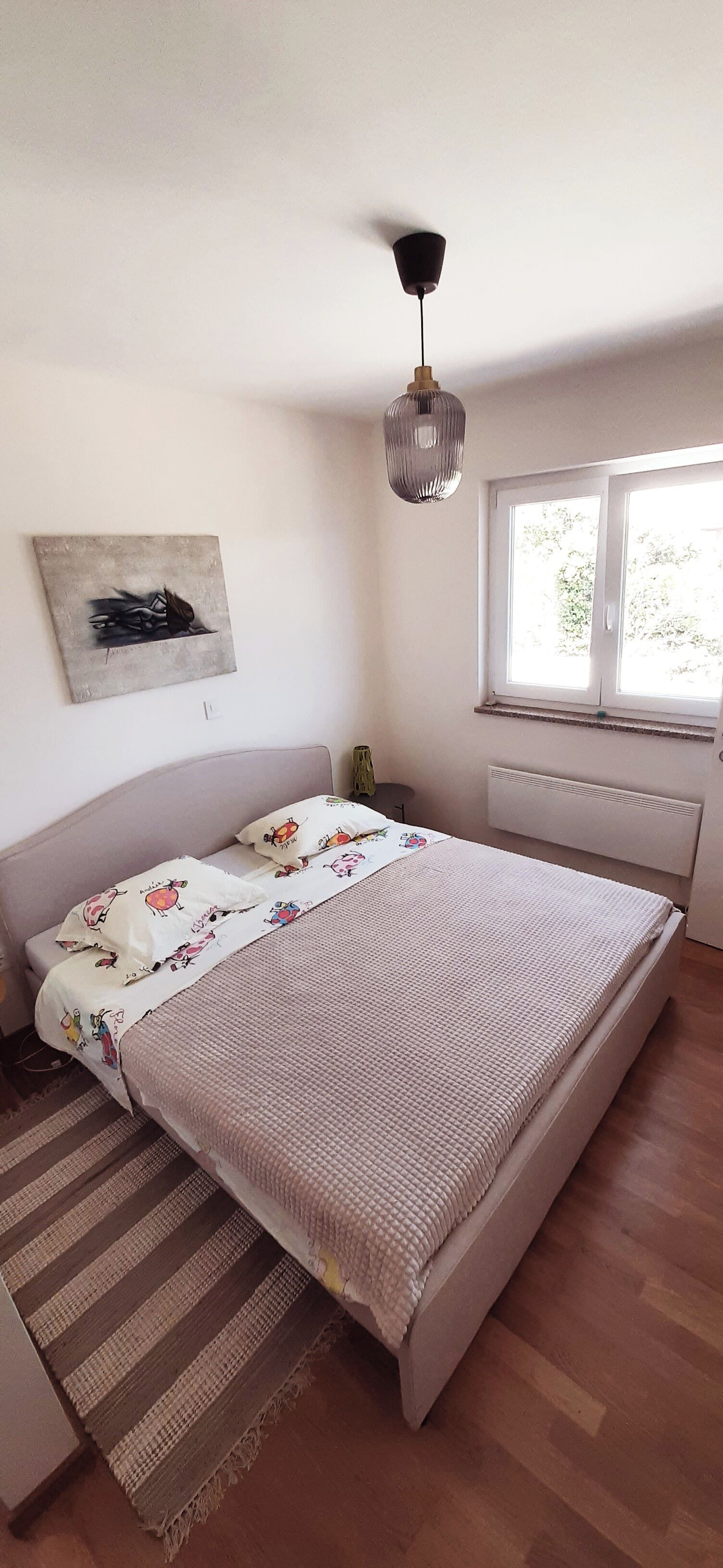 Een slaapkamer met een bed, een raam met zonwering, een radiator, een hanglamp en een schilderij aan de muur.