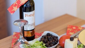 A bottle of Postup wine, a bowl of grapes, a tomato, cheese, and some greens on a wooden table.