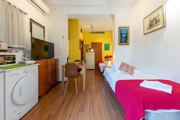 A compact hotel room with a washing machine, a TV, a dining area, and a bed with a red blanket.