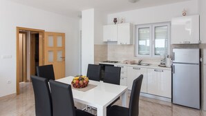 A modern kitchen with white cabinets, a built-in oven, and a refrigerator. There is a dining table with chairs and a fruit bowl.