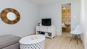 Ein kompakter Wohnraum mit Fernseher auf einem wei ßen Schrank, einem runden weißen Tisch und einem grauen Sofa.
