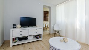 Ein modernes Wohnzimmer mit einem Flachbildfernseher auf einer weißen Konsole, einem runden Couchtisch mit Dekoration und Blick in einen Essbereich mit weißem Tisch und Stühlen.