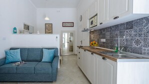 Una cocina compacta con armarios blancos, un microondas y un salpicadero de azulejos. En la zona de estar contigua, hay un sofá azul con cojines.