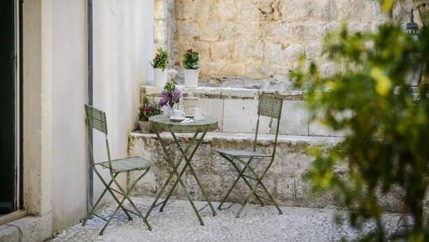 Un pequeño espacio al aire libre con una mesa y dos sillas, una planta en maceta y un muro de piedra al fondo.