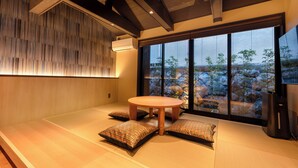 A modern interior with a tatami mat floor, a round wooden table, and a large window offering a view of a garden. - Kanade GionRokuhara (Kyoto)