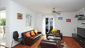 Una sala de estar con un sofá, sillas, una mesa de centro y un televisor. Hay un ventilador de techo, una ventana con cortinas y una puerta que da al exterior.