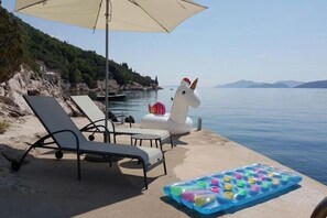 Eine Strandlounge mit Liegestühlen, einem weißen aufblasbaren Einhorn und einer bunten Kühlauflage im Sand.