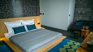 Quarto de hotel moderno com uma cama grande, cabeceira de madeira, carpete estampado e um pequeno espaço de estar com mesa e cadeiras.