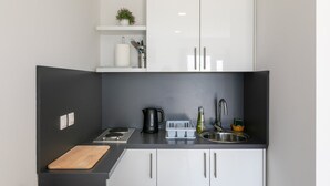 Una cocina compacta con encimera negra, armarios blancos, un lavamanos de acero inoxidable, una tetera y una tabla de cortar.
