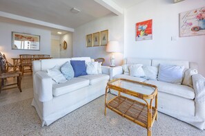 A living room with two white sofas, a glass coffee table, and a dining area with wicker chairs.