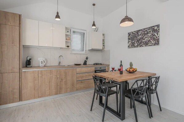 A modern kitchen with wooden cabinets, a wooden dining table, and black chairs. There is a window with blinds, a coffee maker, and a fruit bowl on the table.