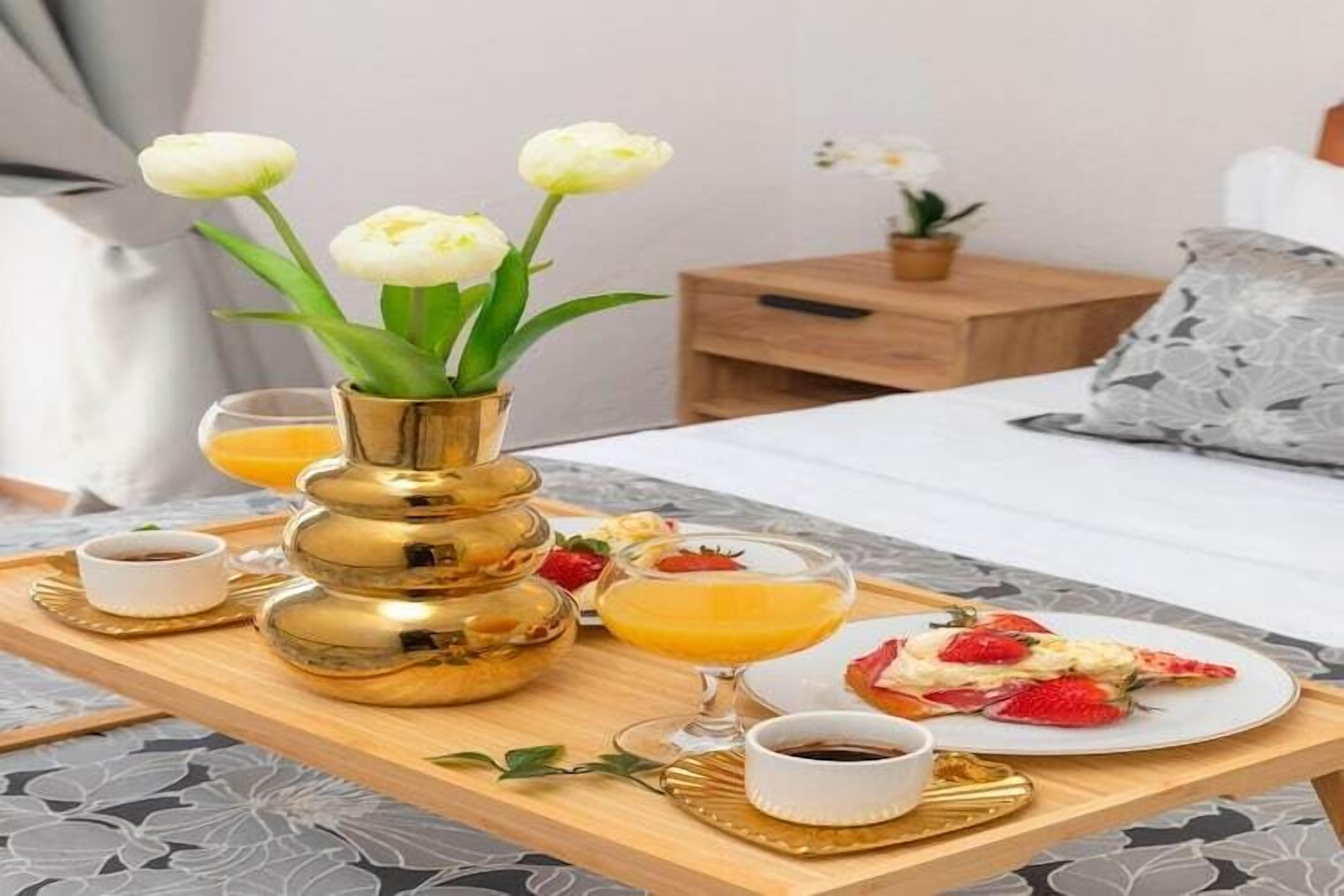 A breakfast tray with a golden vase of white flowers, a plate of food, a glass of orange juice, and a cup of coffee.