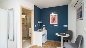 Ein kleines Zimmer mit Dusche, einer kleinen Küchenzeile, einem Essbereich mit Tisch und Stühlen und einem gerahmten Kunstwerk an der Wand.