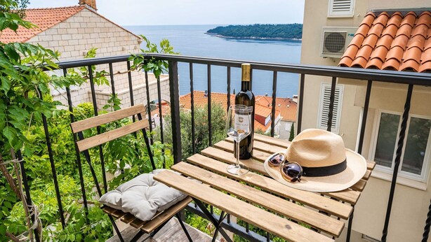 Ein Holztisch mit einer Weinflasche, Gläsern und einem Hut auf einem Balkon mit Blick auf ein Gewässer.