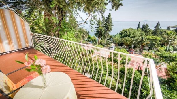A balcony with a view of the sea, surrounded by lush greenery and a white railing.