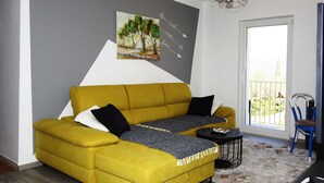 A yellow sofa with a grey throw blanket, a small black table, and a blue chair in a room with a grey wall and a painting. - Apartment Marita - One Bedroom Apartment with Balcony (Cilipi)