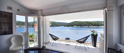 A balcony with a view of a lake and hills, furnished with a white chair, two black chairs, and a potted plant.