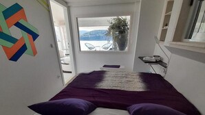 A bedroom with a bed, purple pillows, a window with a view of mountains, and a colorful wall decoration. - Apartment OM - One Bedroom Apartment with Balcony and Sea view (Povlja)