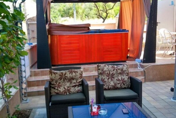 A covered outdoor seating area with two wicker chairs and a glass table, a red door in the background, and a small plant to the left.