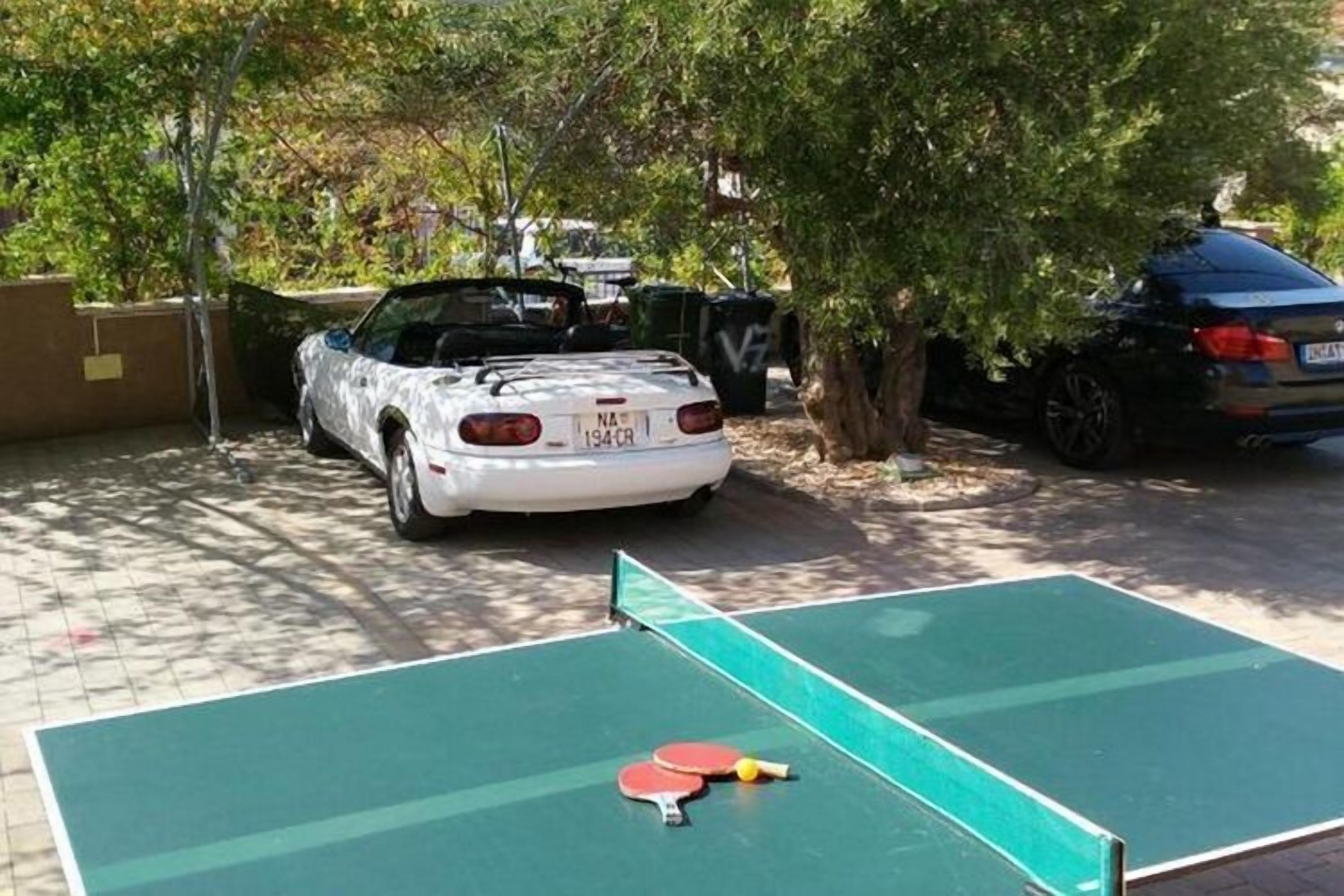 Una mesa de ping pong con dos paletas y una pelota, un coche blanco aparcado detrás y otro negro a la derecha.