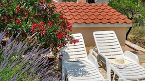 Une terrasse avec des chaises longues en plastique blanc, un petit bâtiment doté d’un toit en tuiles et des plantes de lavande tout autour.