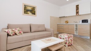 Una sala de estar moderna con un sofá color beige, una mesa de centro blanca y un reposapiés estampado. Junto a la sala, una cocina con microondas, un fregadero y un armario de madera.