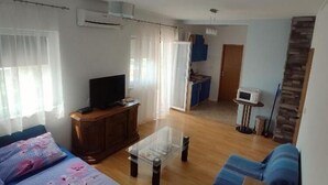 Ein Wohnzimmer mit einer Couch, einem Couchtisch und einem Fernseher. Im Hintergrund ist eine Küchenzeile erkennbar.