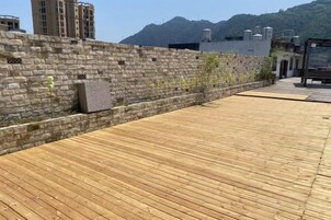 Una terraza de madera con un muro de piedra, una montaña al fondo y un edificio con tejado plano.