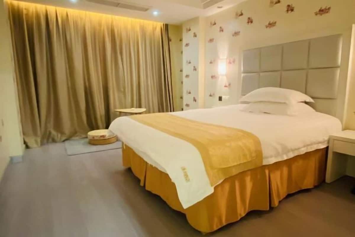 A hotel room with a large bed, a bedside table, a small stool, and a curtain.