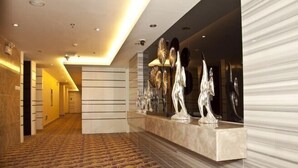 Un hall d’hôtel moderne, avec un tapis à motifs, un comptoir de réception agrémenté de sculptures métalliques décoratives et un couloir menant aux autres chambres.