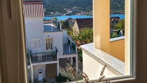 A view from a window overlooking a building with a balcony, a courtyard, and a distant view of a body of water and hills. - Apartments Vala - One Bedroom Apartment with Balcony and Sea View A4 (Vinisce)