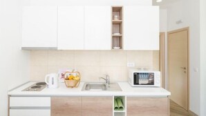 A modern kitchen with white cabinets, a sink, a kettle, a microwave, and a basket of fruit.