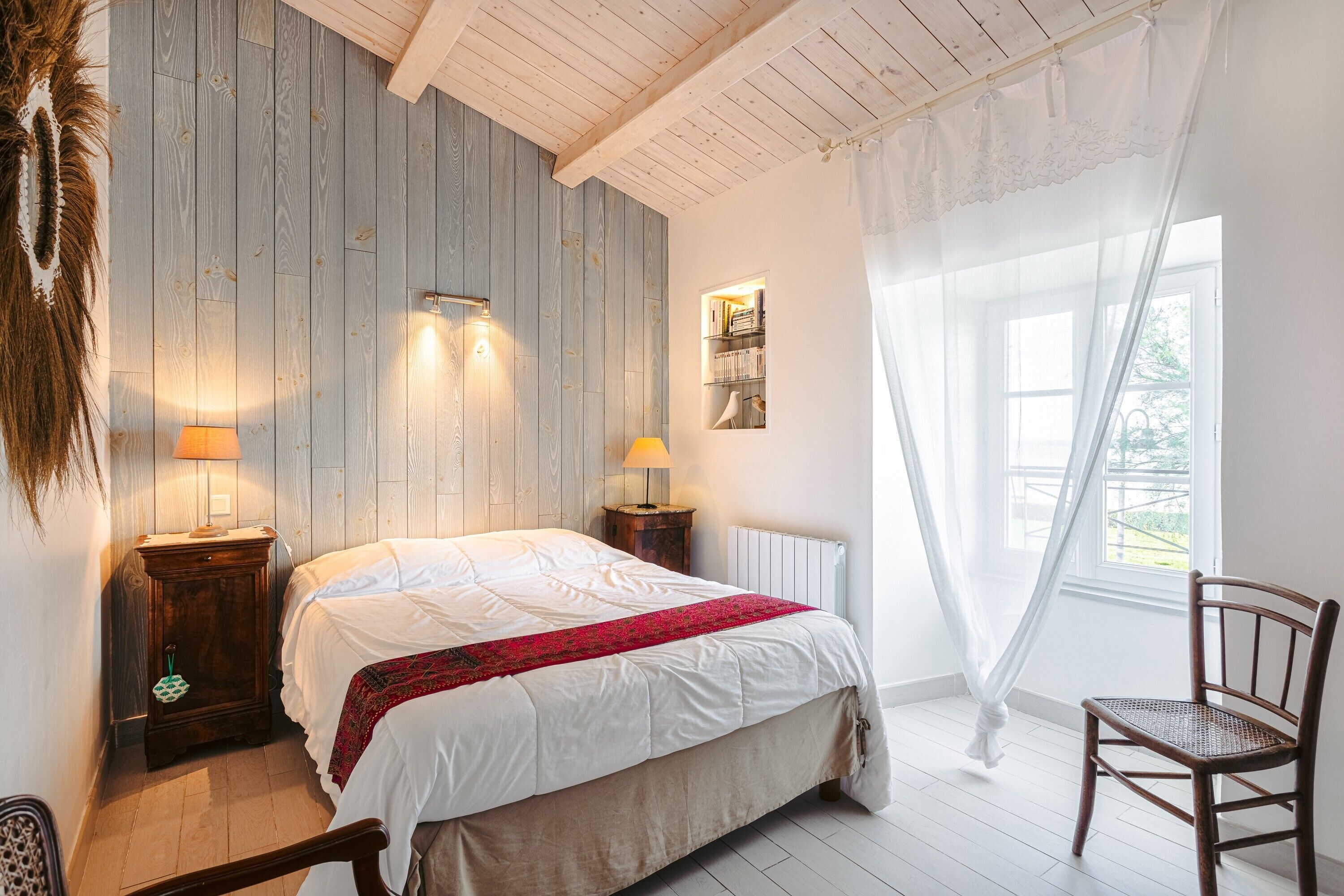 A bedroom with a bed, two bedside lamps, a wooden headboard, a window with sheer curtains, and a radiator.