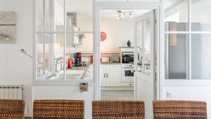 Une cuisine moderne avec des meubles blancs, une horloge rouge et une vue sur le coin repas, où se trouvent des chaises en osier.