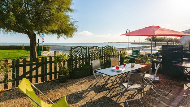Un espace extérieur aménagé avec une table, des chaises et un parasol rouge, donnant sur une étendue d’eau.