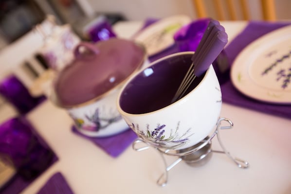 A table with a white tablecloth and purple napkins. There are two white cups with purple floral designs and a purple teapot.