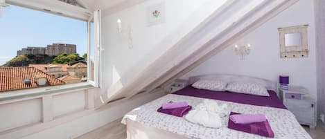 A bedroom with a bed, a nightstand, a window with a view of a castle, and a white ceiling.