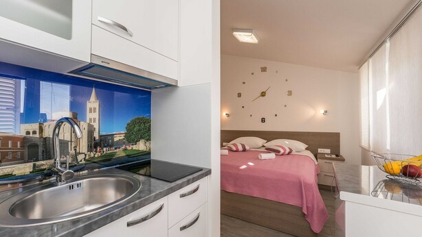 A modern kitchen with a stainless steel sink and a decorative backsplash. The room has a bed with a pink bedspread and a wall clock.