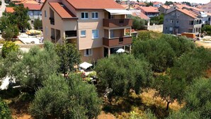 A multi-story building with balconies, surrounded by olive trees and other vegetation.