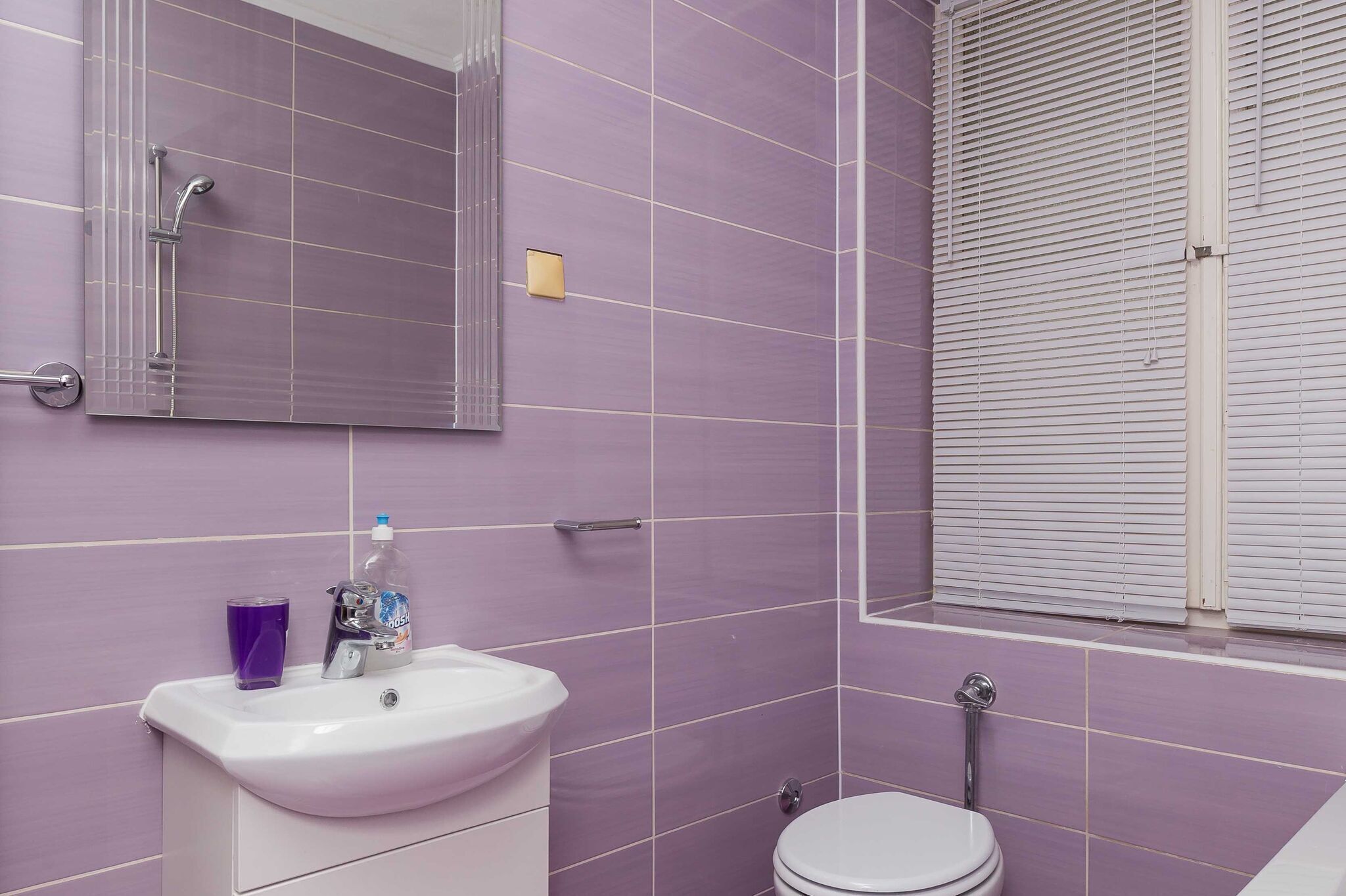 Ein Badezimmer mit violetten Fliesen, einem weißen Waschbecken, einem Spiegel, einem Fenster mit Jalousien und einer Toilette.