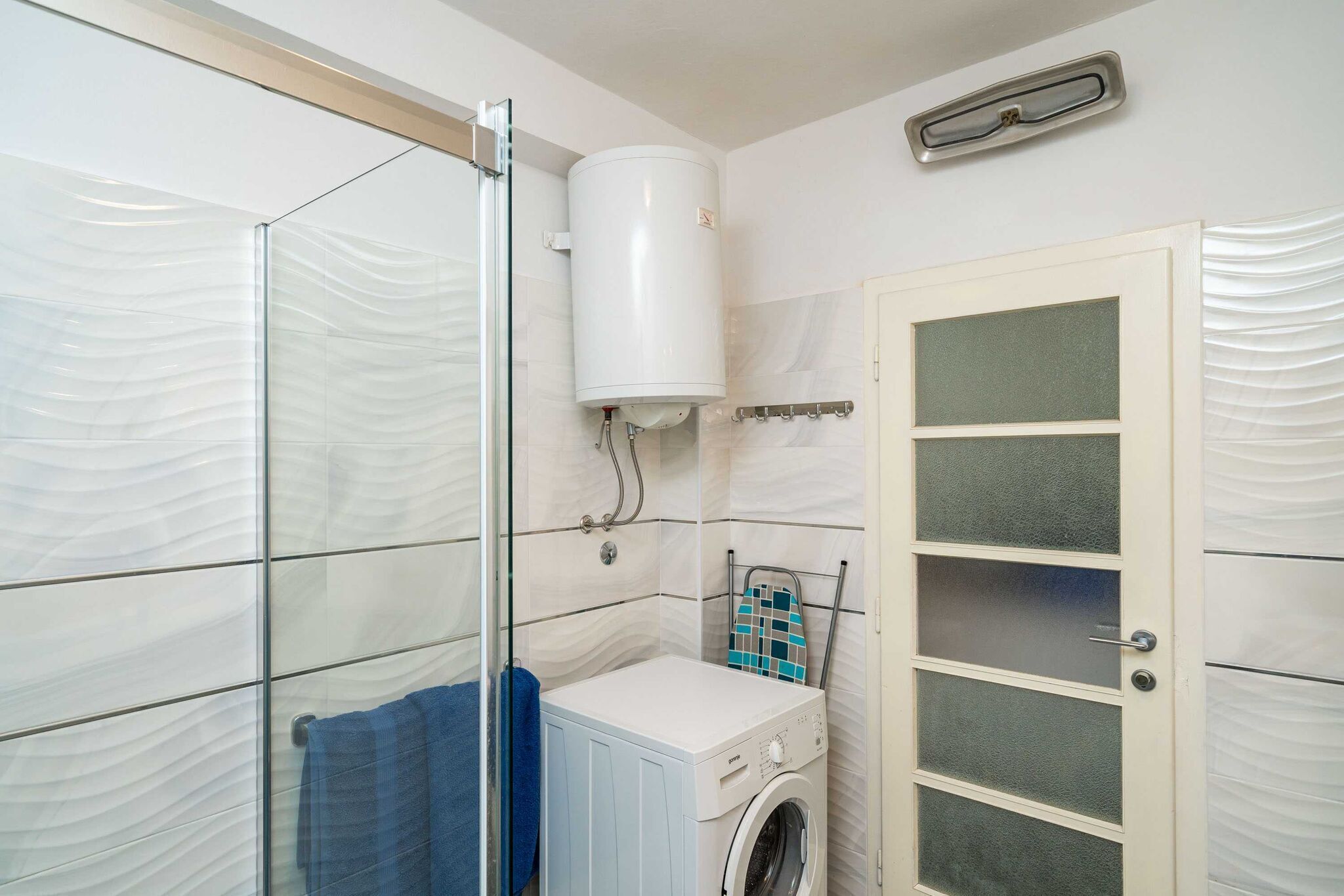 Un baño pequeño con cabina de ducha de vidrio, lavadora integrada y un revestimiento de pared con textura.