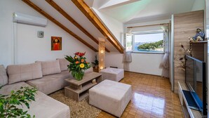 A cozy living room with a wooden ceiling, a beige sofa, a small table with a vase of flowers, and a view of the outdoors through a window. - Apartments & Rooms Mara & Petrunjela - One Bedroom Apartment with Balcony and Sea View (Cavtat)