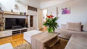 A modern living room with a flat-screen TV, a wooden coffee table, a beige sofa, and a bookshelf. - Apartments & Rooms Mara & Petrunjela - One Bedroom Apartment with Balcony and Sea View (Cavtat)