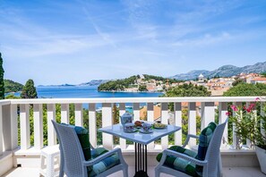 Ein Balkon mit Tisch für zwei Personen, mit Blick auf eine Küstenstadt und freier Sicht auf das Meer und die Berge.