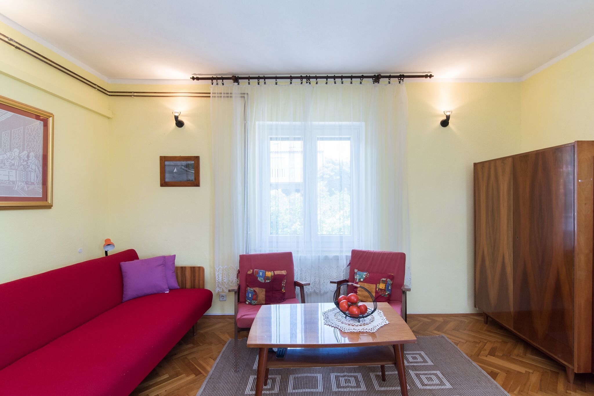 A living room with a red sofa, two armchairs, a wooden table with a fruit bowl, a wooden wardrobe, and a large window with sheer curtains.