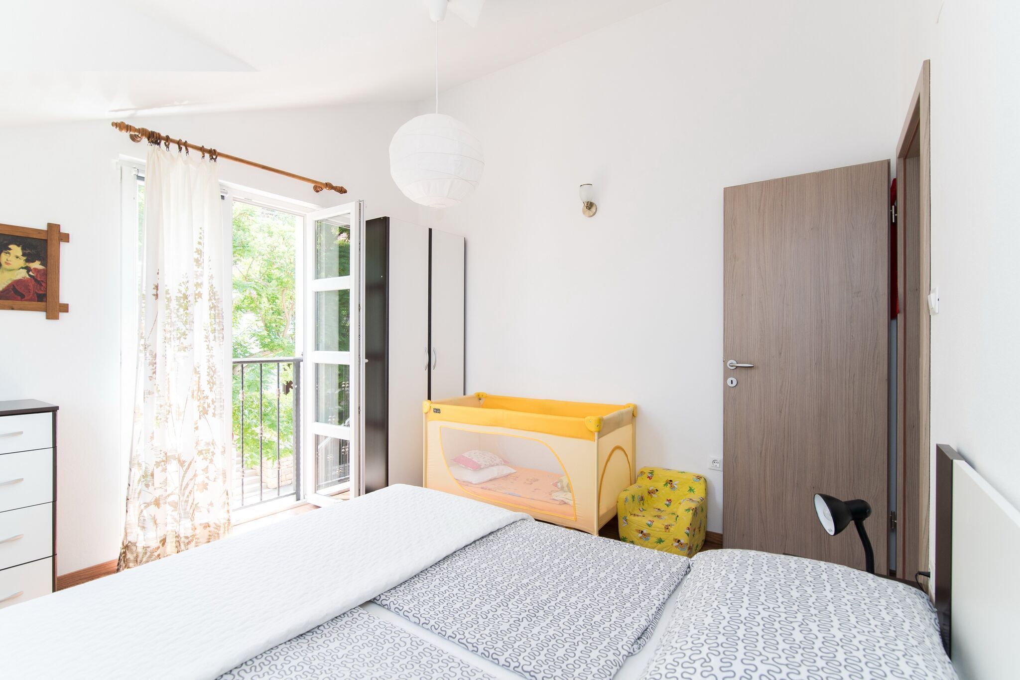 A bedroom with a bed, a window with curtains, a bedside table, a lamp, a wardrobe, and a small playpen.